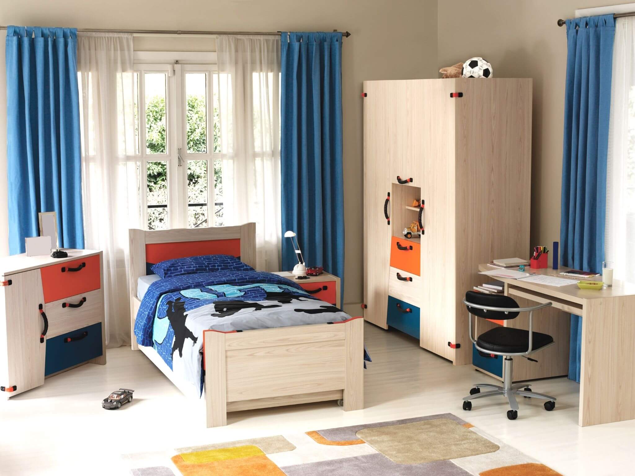 A child's bedroom with blue full length curtains, blue and orange bedding and light oak wardrobe, computer desk and drawers.