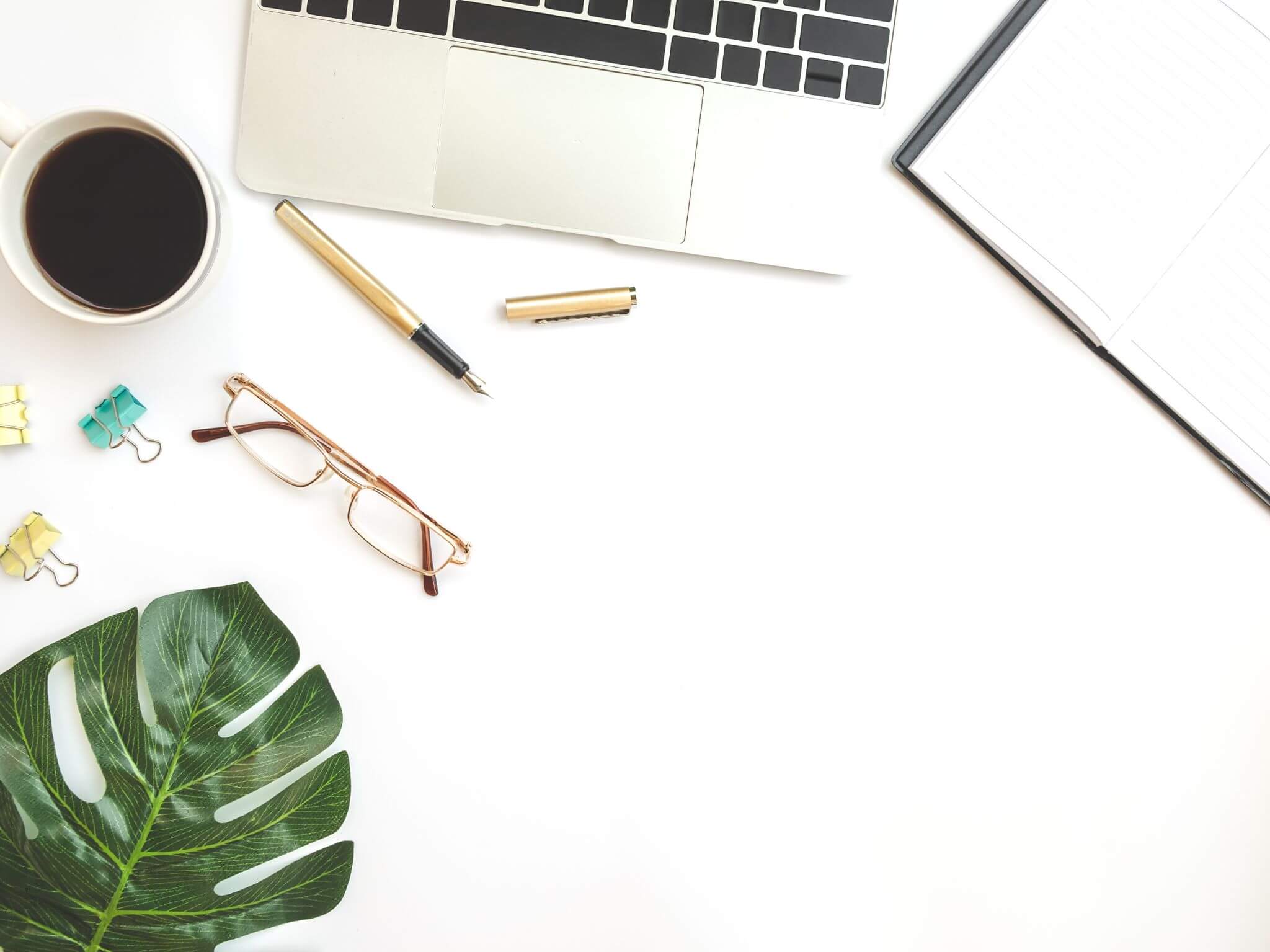 A white desk top wth an open laptop. a mug of black coffee and a pair of glasses.