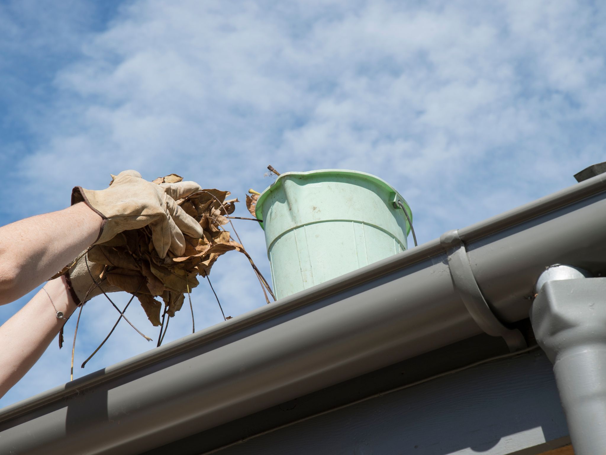 Hands clearing fallen leaves from a gutter with a green bucket in the background
