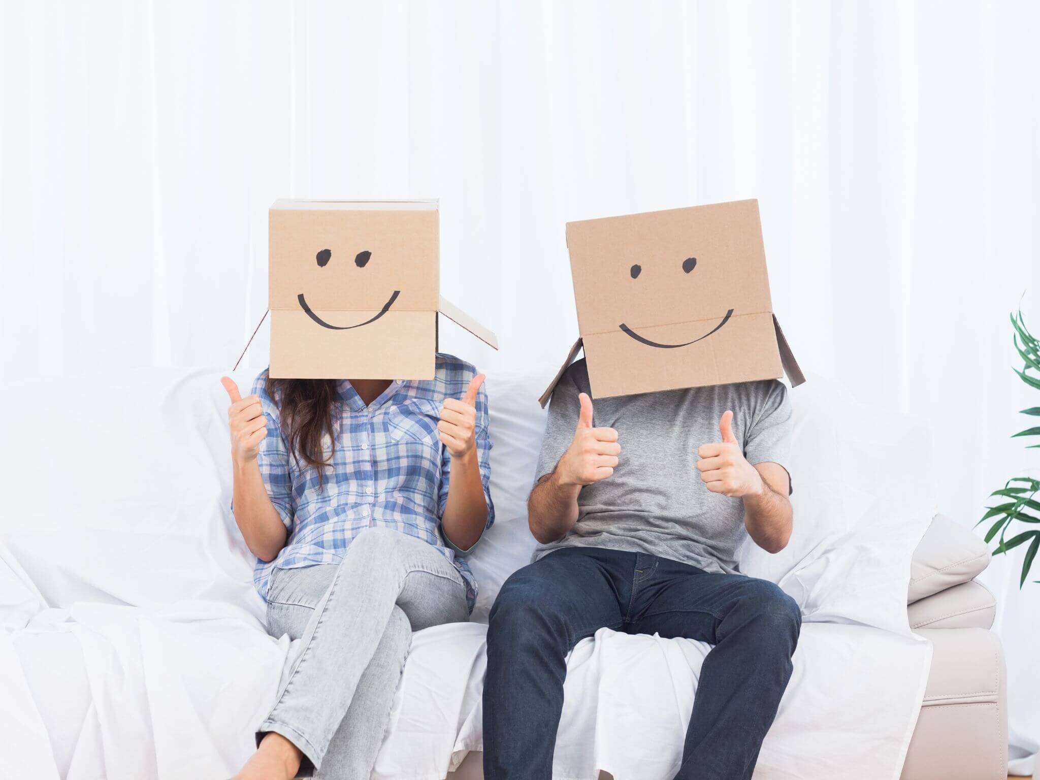 A male and female couple sitting on a sofa with cardboard boxes on their head whilst holding their thumbs up. The boxes have smiley faces drawn on the front.