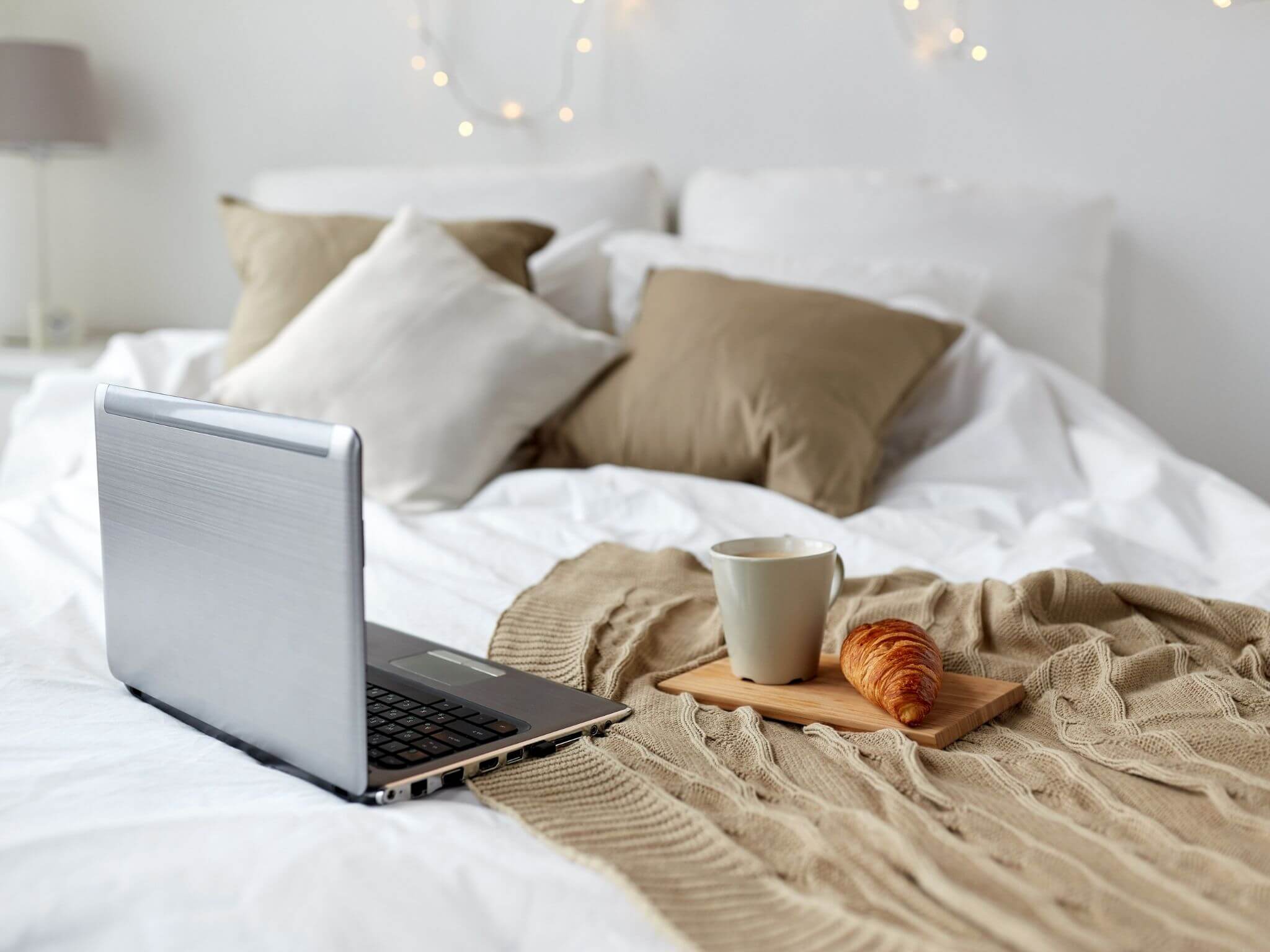 A cosy bed with beigh and cream scatter cushions with a laptop and croissants and a cup of tea on a tray.
